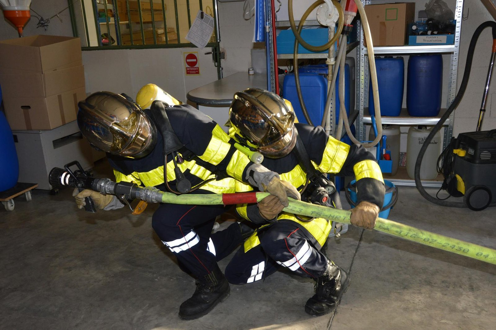 Two firefighters in full gear coordinate to handle a fire hose inside a warehouse setting.